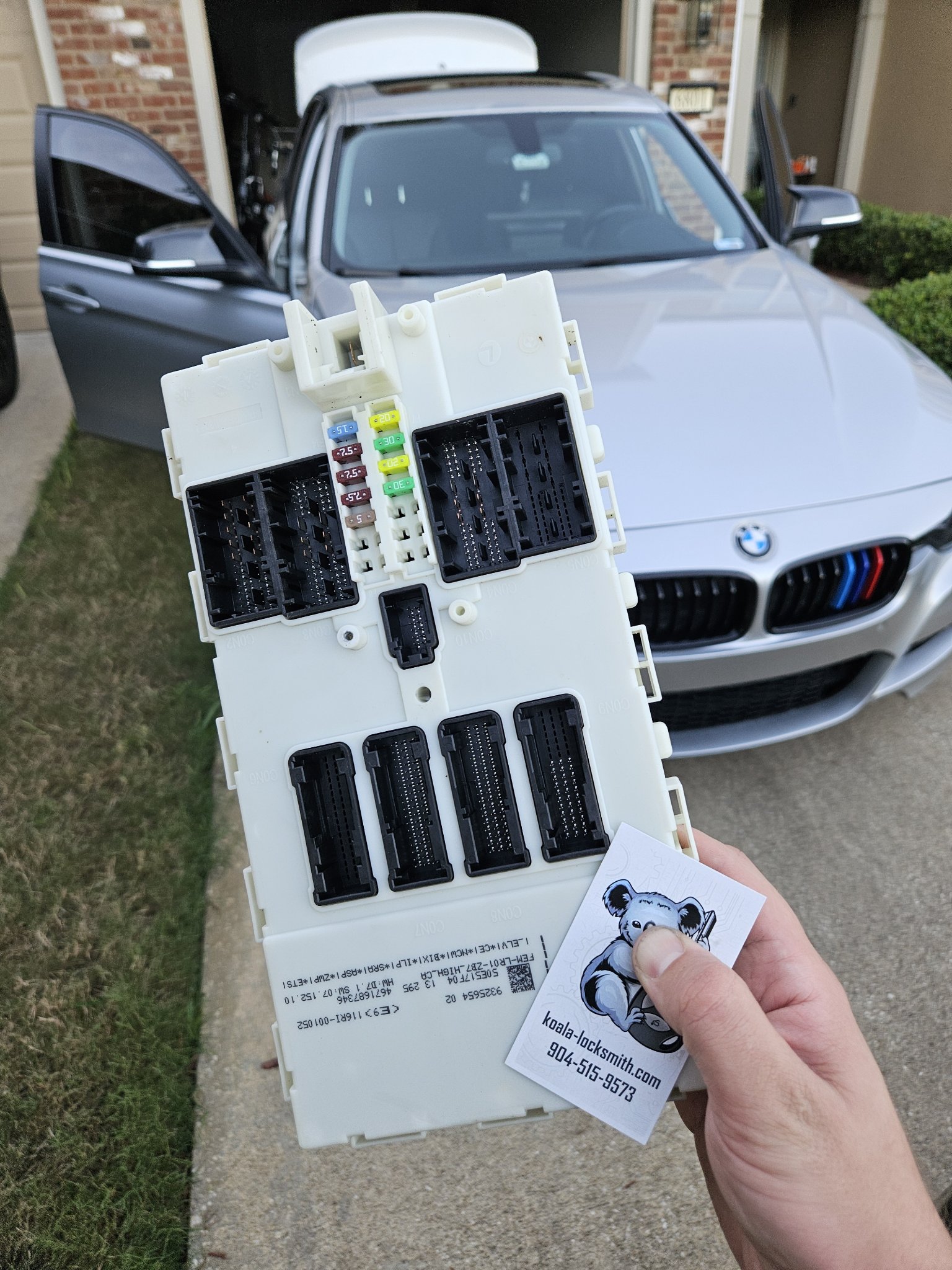 Removed BMW module held in front of the silver 320i with a Koala Locksmith card